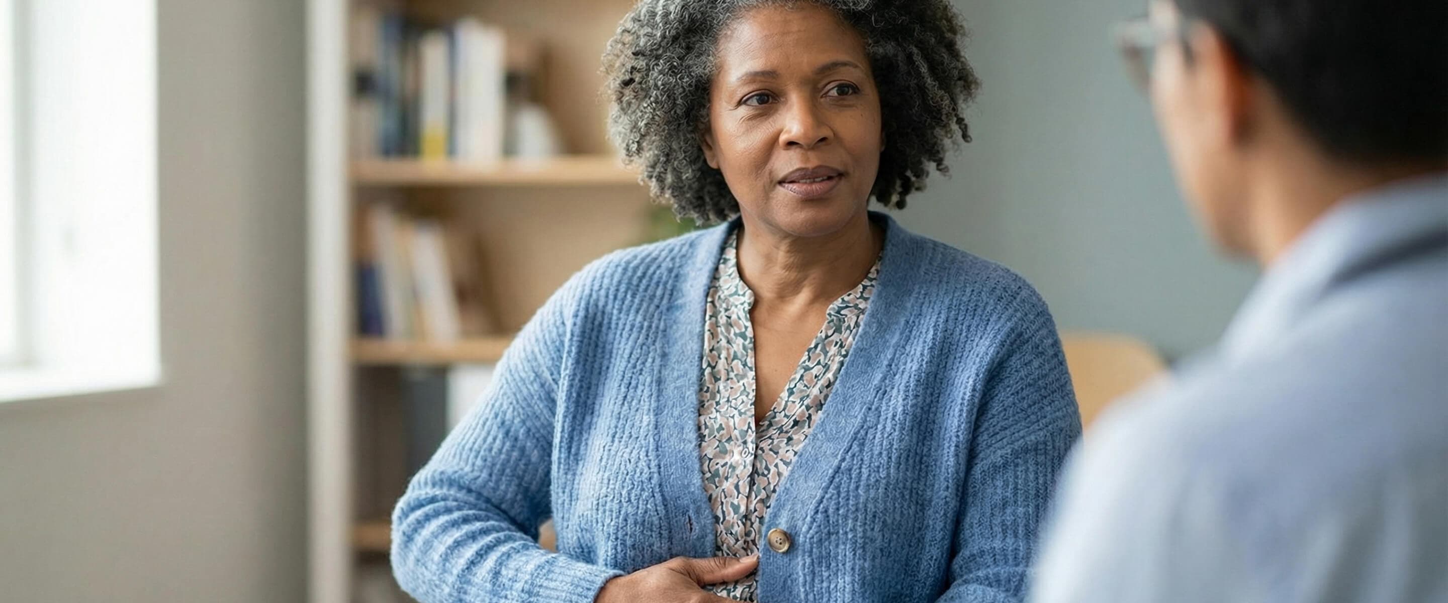 A woman in a blue cardigan sits and talks with another person in an office setting, with shelves in the background.